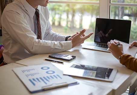 Two people sit at a desk with documents, a calculator, a tablet, and a laptop, discussing business or financial reports near a window with greenery outside. One person gestures while the other takes notes.