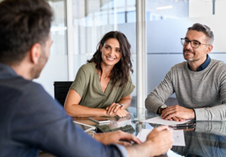 Three people sit at a glass table in a modern office, engaged in a discussion. A woman and a man face another man whose back is to the camera. They all appear to be smiling and attentive.