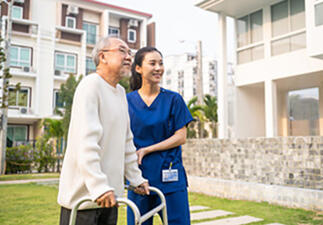 An elderly man using a walker is assisted outdoors by a smiling female caregiver in blue scrubs. They are standing in front of a modern residential building on a sunny day.