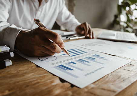 A person in a white shirt reviews business charts and graphs on printed papers at a wooden desk, using a pen to point at a bar graph, with a laptop and documents nearby.