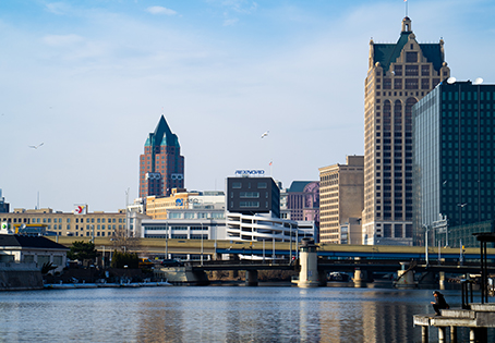 Milwaukee city skyline with tall buildings, including the Milwaukee Center, viewed from across the river under a blue sky, with a bridge spanning the water in the foreground.