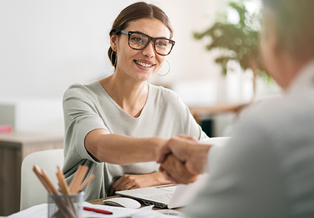 A woman wearing glasses and a light-colored top is smiling and shaking hands with another person across a desk, suggesting a positive or successful meeting. A laptop and pencil holder are visible on the desk.