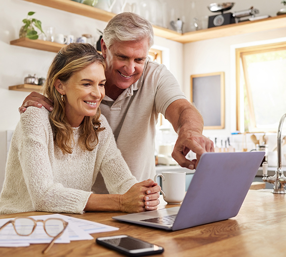 Smiling older couple sits at a kitchen table, looking at a laptop together. The man points at the screen while the woman leans in, both appearing happy and engaged. Papers, a phone, and glasses are on the table.