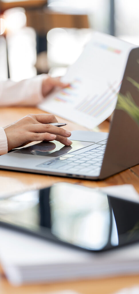 A person works on a laptop while holding a sheet with colorful graphs. A tablet and some documents are on the wooden table, suggesting a work or study environment.