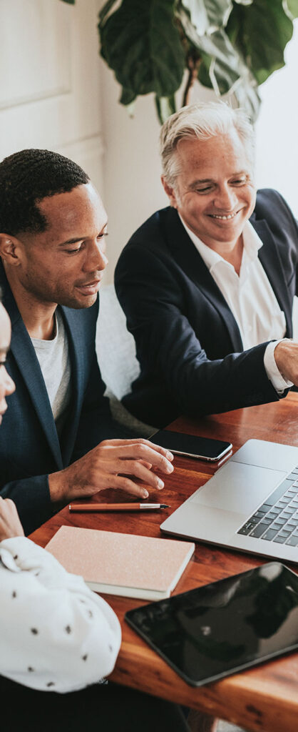 Two men in business attire sit at a table, smiling and working together on a laptop. Notebooks, a smartphone, and a tablet are on the table. A large plant is in the background.