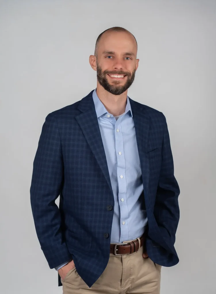 A man with a beard and short hair is smiling while wearing a blue checked blazer, light blue shirt, and khaki pants. He has one hand in his pocket and stands against a plain light background.