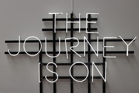 A white neon sign reads THE JOURNEY IS ON against a gray wall with a black grid-patterned background.